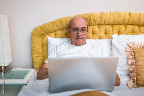 Content senior man with eyeglasses in pajamas enjoying working on laptop while sitting on cozy bed at his home.