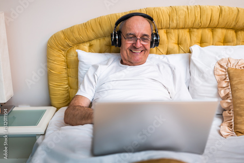Happy senior man in pajamas wearing wireless headphones and working on laptop while sitting on bed in his cozy bedroom.