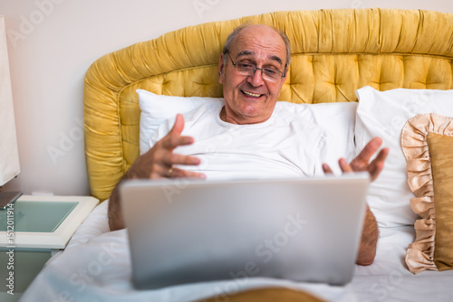 Cheerful senior man in pajamas using laptop while sitting in bed. He is talking and smiling during video call from the comfort of his home. 