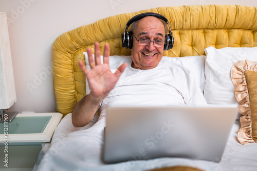 Joyful senior man in pajamas using laptop and wireless headphones after waking up in bed in the morning. He is smiling and waving during a video call from his bedroom.