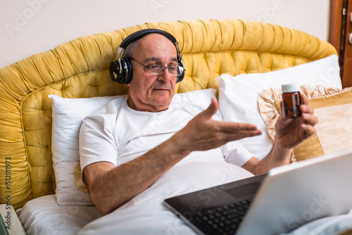 Senior man with headphones holding pill bottle and talking with doctor on laptop while lying in bed. He is having medical consultation via video call from his home.