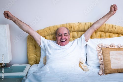 Smiling senior man stretching in bed after waking up at dawn.