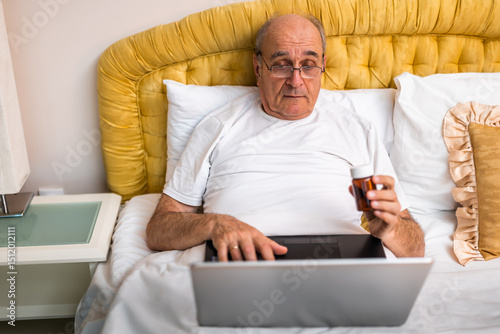 Senior man holding pill bottle and analyzing medical information on Internet via laptop while sitting on bed in his bedroom. 