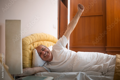 Happy senior man stretching in bed after waking up in early morning hours.