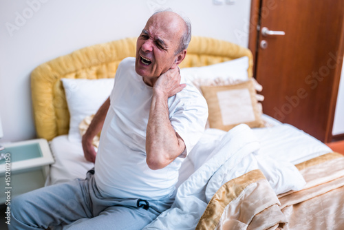 Senior man having pain in neck, he is shouting while sitting on the bed after waking up in the early morning hours.