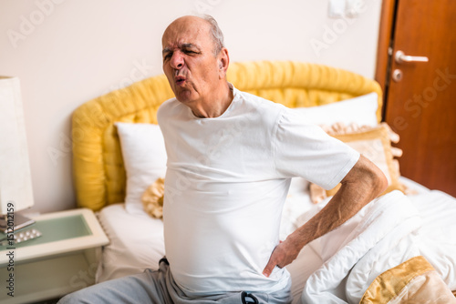 Senior man having pain in back after waking up and sitting on bed in the morning.