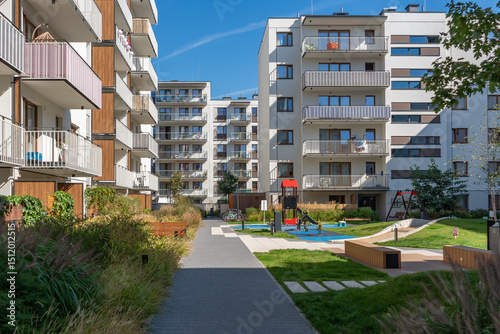 Multifunctional community space in a new residential development. The landscaped courtyard features a children's playground, wooden benches, a green lawn, and a pedestrian walkway.