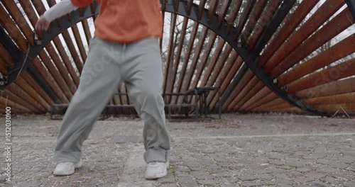 Young woman dancing contemporary or modern dance choreography in an urban park setting. She's wearing baggy gray pants and an orange hooded sweater.