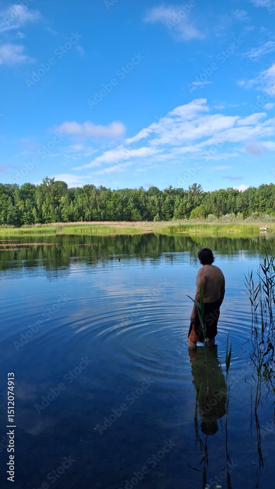 Fototapeta premium Tranquil lake scene with clear blue water, green reeds, and a lush forest under a bright sky. Perfect nature landscape for relaxation, travel, or outdoor themes.