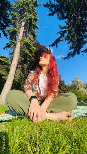Woman sitting on grass in park under blue sky enjoying sunny day with trees surrounding her