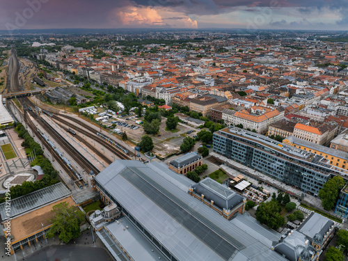 Fototapeta Naklejka Na Ścianę i Meble -  Aerial view of Budapest, Hungary, featuring a large railway station with a glass and metal roof, train tracks, historic buildings, and stormy skies.