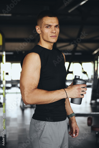 Obraz na plátně Strong and focused male athlete in gym holds a sports shaker with serious expression, reflecting discipline, fitness mindset, and dedication to personal health goals