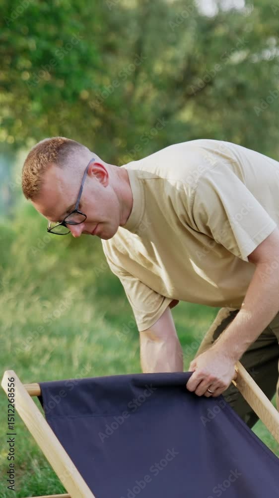 Man in glasses having troubles with unfolding the chair. Finally man succeeds and sits down in a chair. Nature backdrop. Vertical video