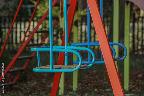 A playground swing set with colorful metal frames, including blue seats and red supports. The vibrant hues and simple design suggest a welcoming play area