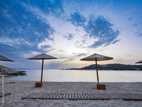 Fototapeta Naklejka Na Ścianę i Meble -  Tranquil beach at sunset with straw umbrellas overlooking the calm water and distant hills in Toroni on Sithonia peninsula, Greece
