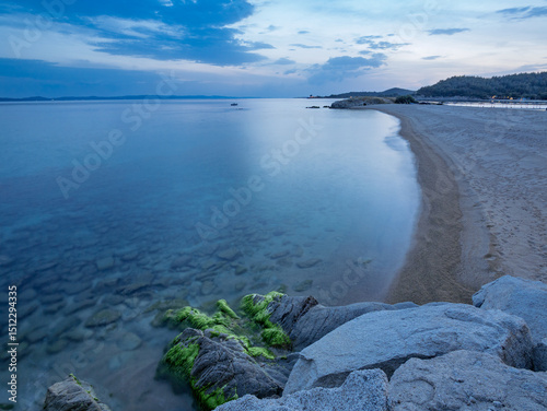 Fototapeta Naklejka Na Ścianę i Meble -  Calm evening at a coastal beach with smooth waves and rocky shoreline under a cloudy sky in Toroni on Sithonia peninsula, Greece