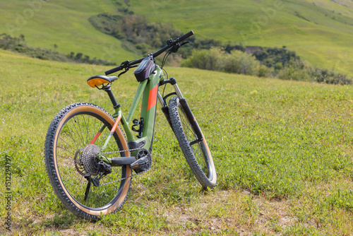 A mountain e-bike is leaned against the ground in a lush green field. Rolling hills stretch in the background under a clear sky, showcasing a peaceful outdoor environment.