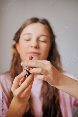 Cute girl applying lipstick with joyful focus in a bright, cozy indoor setting during daylight hours
