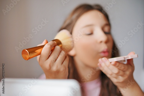 Cute girl applying makeup with a brush while looking into a compact mirror in a cozy indoor setting during the day