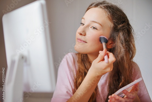 Cute girl applying blush while looking in a mirror during a makeup session at home in the afternoon