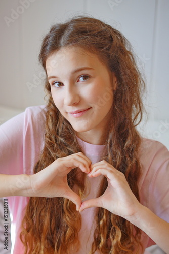 Cute girl with long hair making a heart shape with her hands while smiling warmly indoors on a cozy day