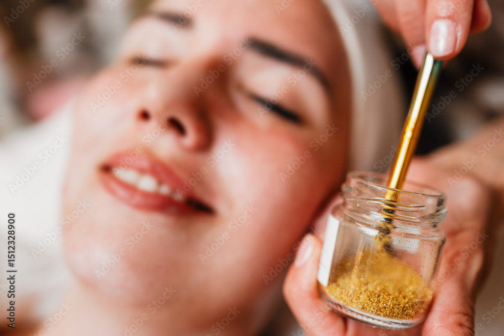 Fototapeta premium Beautician carefully applying gold particles with a brush on the forehead of a woman lying with closed eyes, during a rejuvenating treatment in a beauty spa, enhancing skincare and relaxation