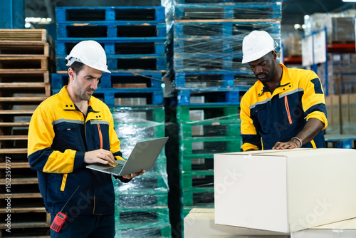Warehouse teamwork between african adult male and caucasian adult male workers wearing uniform and helmet during packing and checking parcels using laptop for shipment preparation process