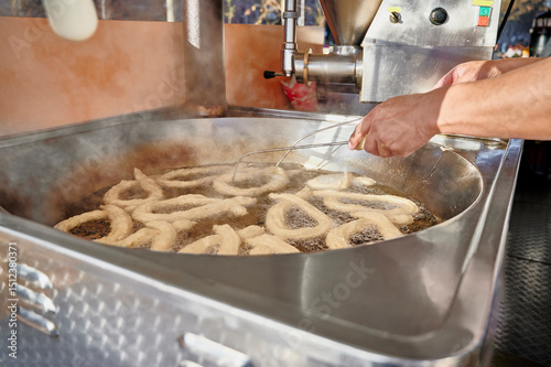 Hands of a churro vendor flipping porras with metal rods in boiling oil inside an industrial deep fryer. Traditional skills and tools used in Spanish churro-making
