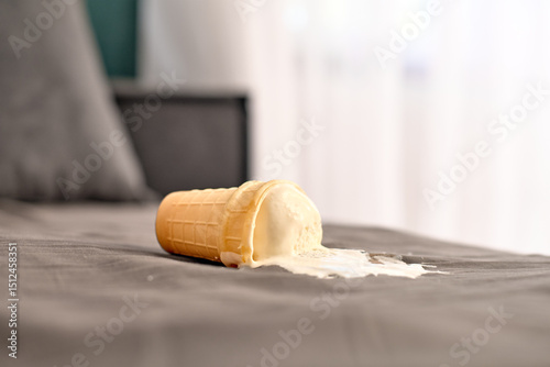 Ice cream cone tipped over on gray bed, ice cream melting into fabric. Bright natural light enters through window creating serene atmosphere. Close-up captures moment of carelessness
