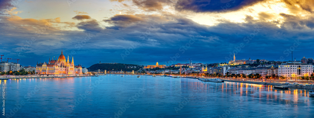 Naklejka premium A panoramic view of Budapest, Hungary, featuring the illuminated Hungarian Parliament Building and Buda Castle with the Széchenyi Chain Bridge spanning the Danube River under a vibrant evening sky