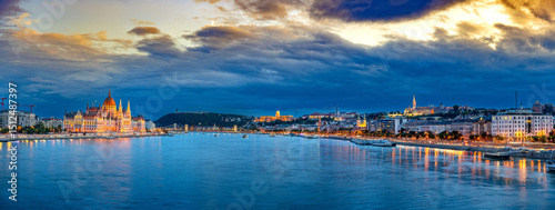 A panoramic view of Budapest, Hungary, featuring the illuminated Hungarian Parliament Building and Buda Castle with the Széchenyi Chain Bridge spanning the Danube River under a vibrant evening sky