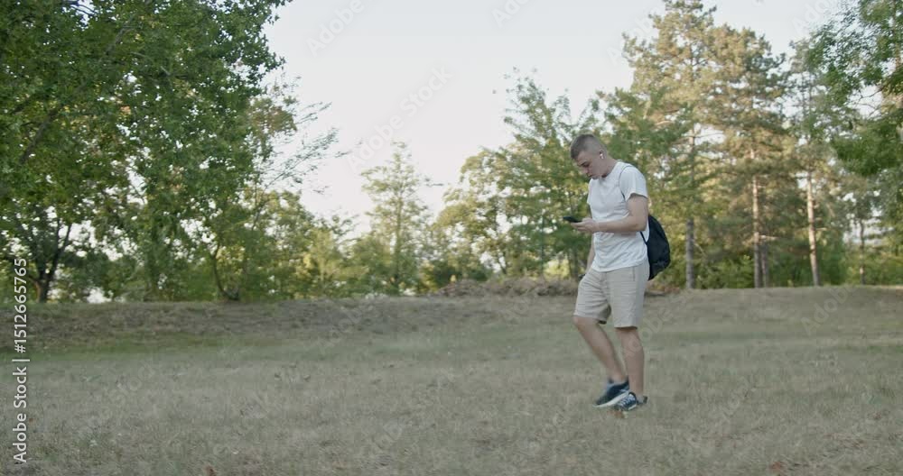 A young man walks through a park while looking at his phone and carrying a water bottle. He is wearing casual clothes and has a backpack. A young man walks through a park while looking at his phone and carrying a water bottle. He is wearing casual clothes and has a backpack.