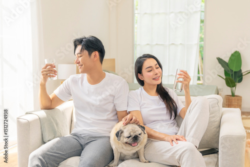 Happy Asian family couple drinking a glass of pure water and playing with senior Pug dog in living room. Man and woman relaxing with domestic dog at home. People healthcare and Pet parent concept.