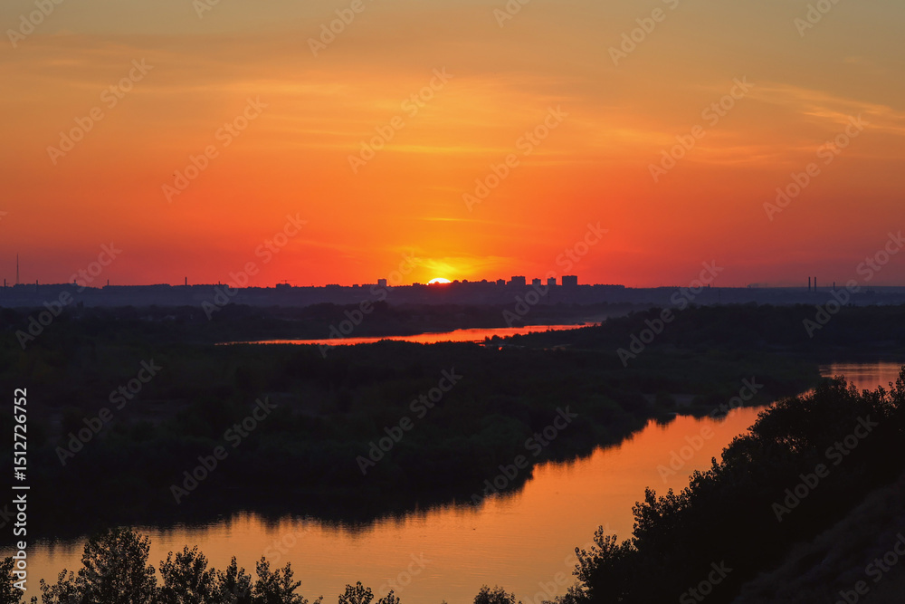 Fototapeta premium The setting sun paints the sky with fiery hues of orange and red, casting a warm glow over the silhouette of a cityscape and a tranquil river meandering through lush vegetation