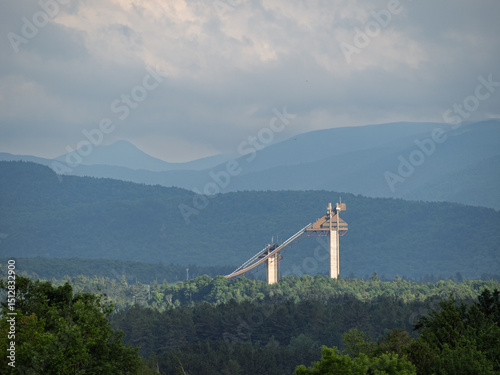 Φωτογραφία The Olympic Jumping Complex in Lake Placid, Upstate New York, lit by late evening sun rays, seen from afar with mountains in the background