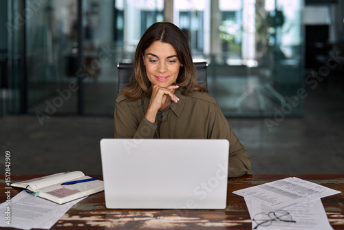 Smiling busy mature middle aged professional business woman, female company manager executive or entrepreneur looking at laptop computer in office working on online financial data sitting at desk.