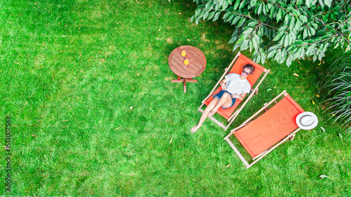 Young girl relaxes in summer garden in sunbed deckchair on grass, woman reading book outdoors in green park on weekend, aerial drone view from above