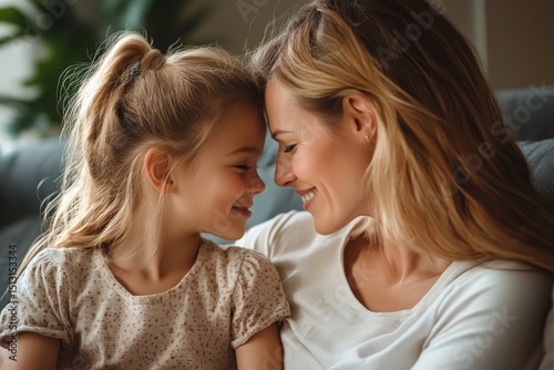 Side view shot of loving mother spending time with her daughter, touching foreheads while sitting on sofa at home, copy space