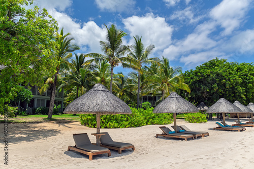 Fototapeta Naklejka Na Ścianę i Meble -  Empty Beach Chairs and Straw Parasols on White Sand by Palm Trees.Luxury Resort Vacation.Relaxing Getaway.Sunbeds with Towels under Thatched Umbrellas on a Sunny Tropical Beach.