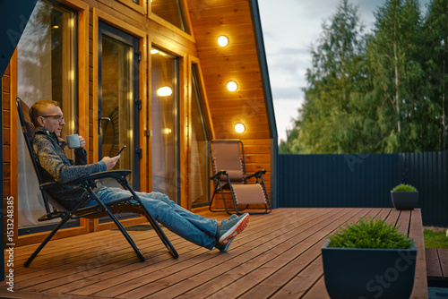 Man sits with a cup of tea in the evening on the terrace illuminated by outdoor wall lamps