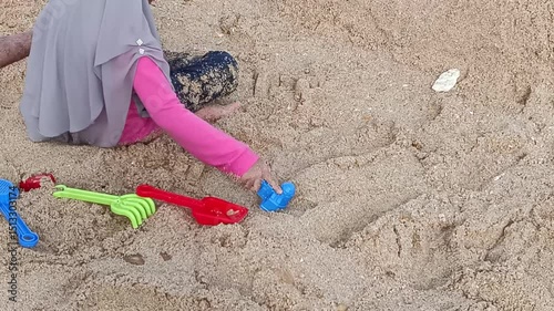 A young child, wearing a headscarf, happily plays with colorful plastic toys in the sand on the beach. She immerses herself in building, exploring, and having fun outdoor.