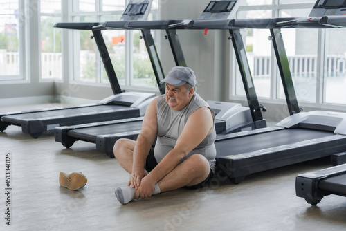 Fat man sits with his hand on his ankle after injuring his ankle while running on a treadmill in a gym.