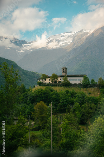 Lonely stone church on a green hilltop surrounded by alpine forest, under dramatic cloudy sky and snowy mountain peaks in Lombardy, Italy.. photo