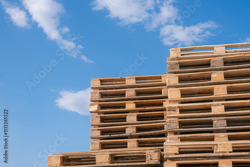 Stacked wooden pallets against blue sky. Large stack of wooden pallets arranged in an orderly manner. Related concept to logistics or warehousing.
