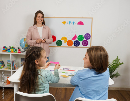 Teacher points at the board with colorful fractions while two students hold up math answer cards. An engaging classroom moment focused on visual learning and active participation.