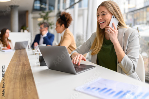 Businesswoman working on laptop and speaking on smart phone with professional colleagues collaborating in the background
