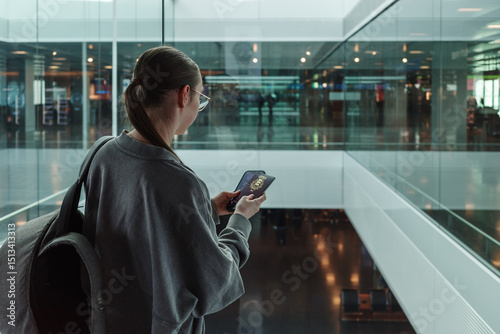 Young woman with a backpack holding a passport and smartphone while looking through glass at a contemporary airport. Concept of international travel, identity verification and global mobility.