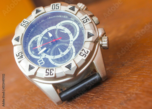 Luxury analog wristwatch with a black leather strap and stainless steel bezel, placed on a wooden surface, showcasing bold numerals and a modern chronograph dial.