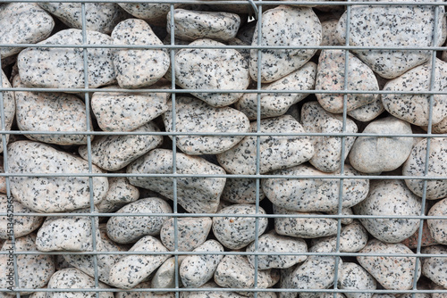  Close-up of rounded granite stones enclosed in a metal gabion cage for landscape construction. Concept of durability, architectural texture and natural stone arrangement in modern design.