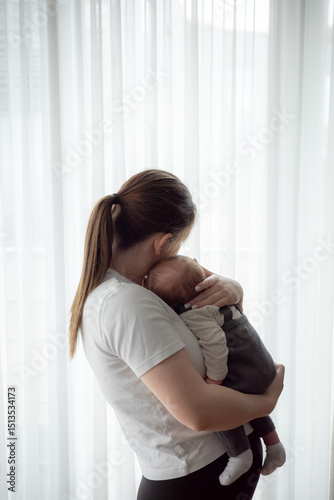 Young mother holding newborn baby in soft natural light standing by a window. Concept of early motherhood, tenderness, bonding, parental love, and infant care in a calm home environment.